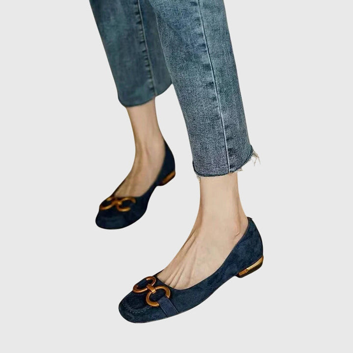 A pair of black slip-on flats with a polished gold ring detail across the toe.