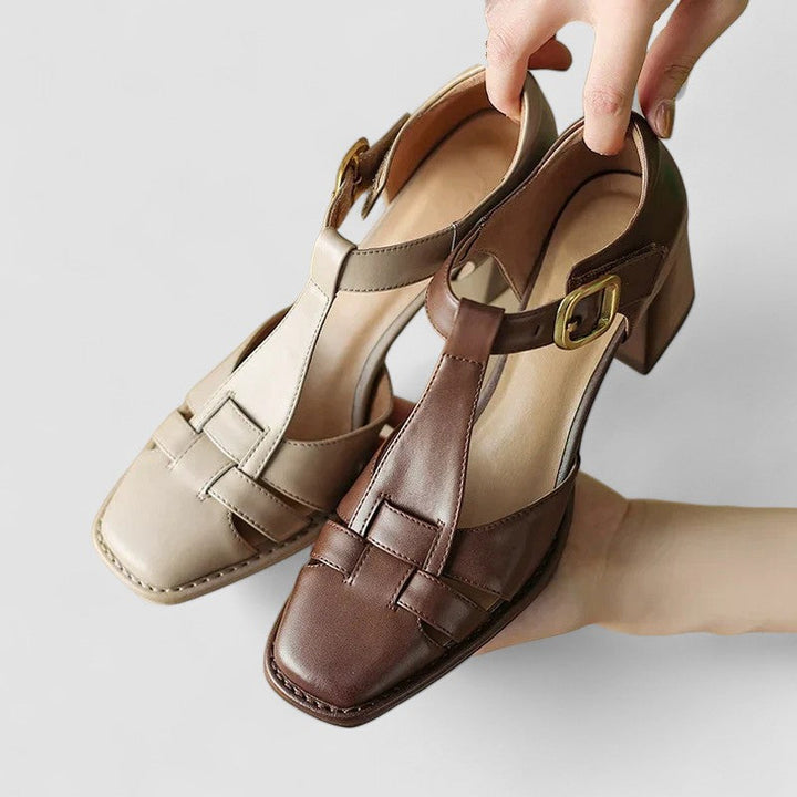 A woman wearing beige t-strap block heel sandals with an adjustable buckle, standing comfortably.