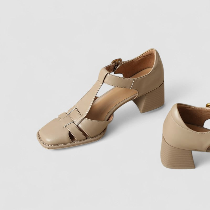 A woman wearing beige t-strap block heel sandals with an adjustable buckle, standing comfortably.