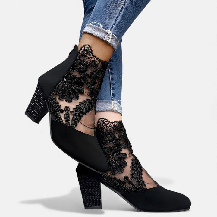 A woman wearing wine block-heel shoes with a sheer black floral-lace ankle overlay and a crystal-set heel