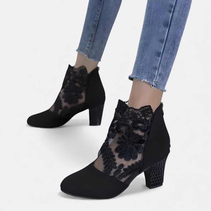 A woman wearing wine block-heel shoes with a sheer black floral-lace ankle overlay and a crystal-set heel