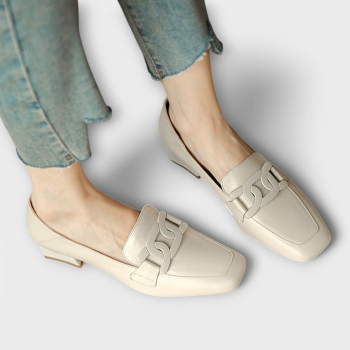 A pair of soft beige square-toe loafers with a chain-link ornament across the front.