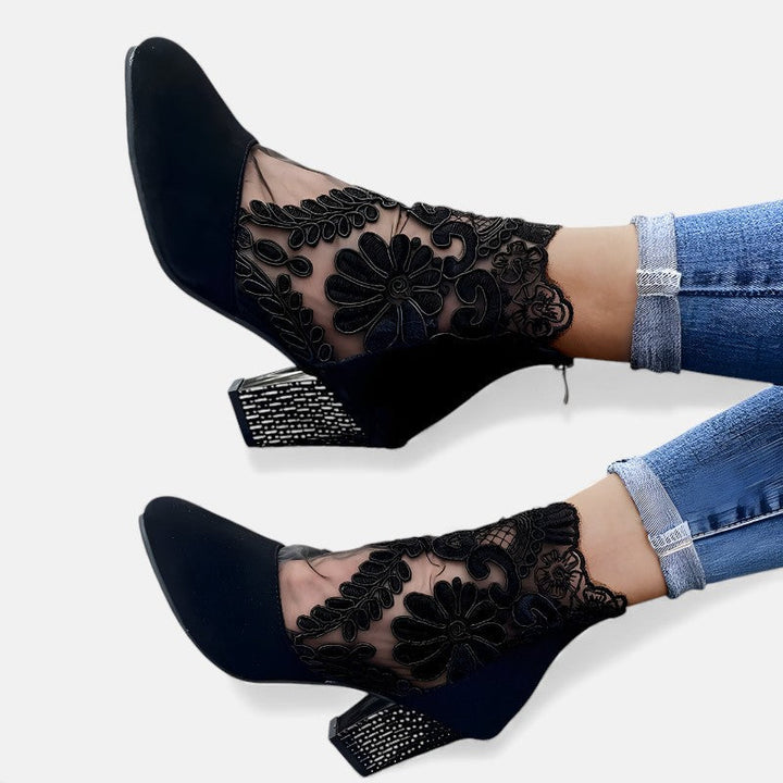 A woman wearing wine block-heel shoes with a sheer black floral-lace ankle overlay and a crystal-set heel