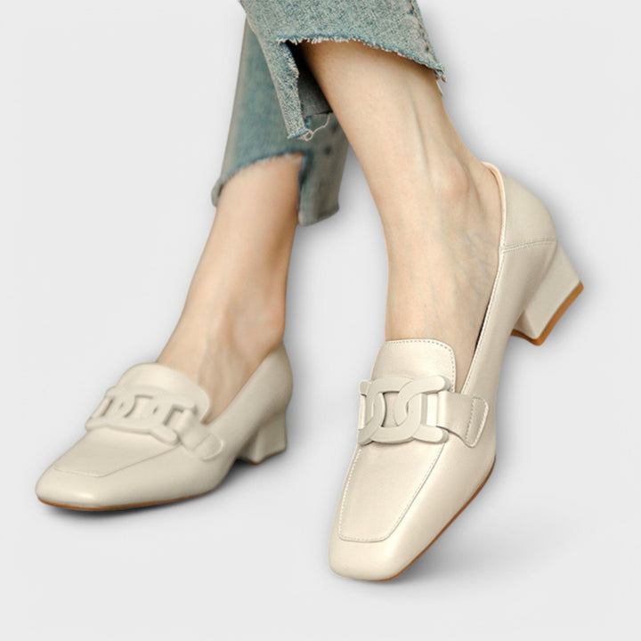A pair of soft beige square-toe loafers with a chain-link ornament across the front.