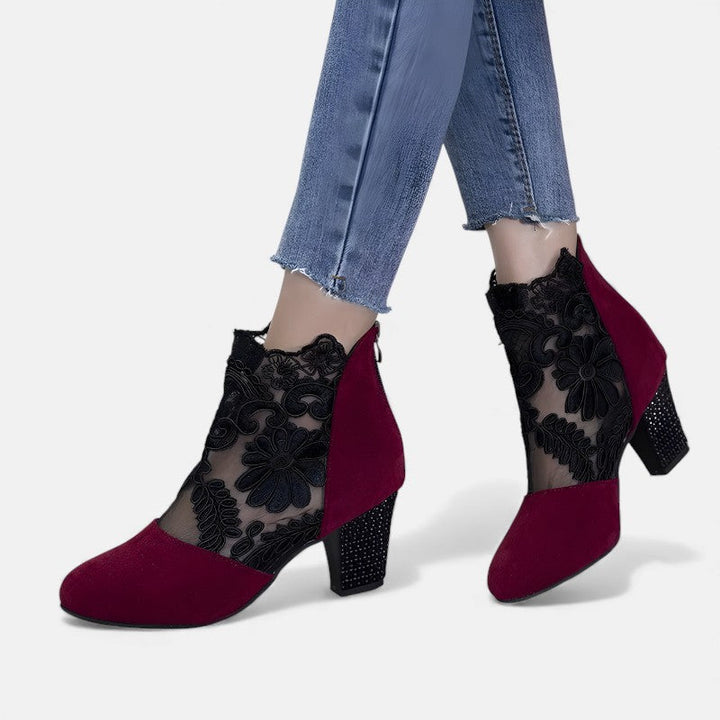 A woman wearing wine block-heel shoes with a sheer black floral-lace ankle overlay and a crystal-set heel