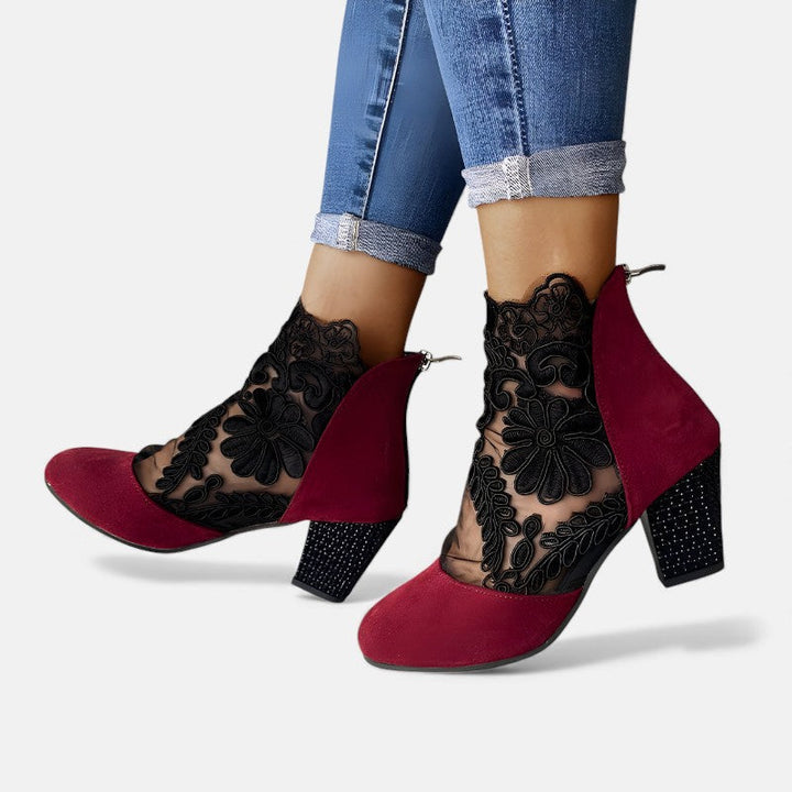 A woman wearing wine block-heel shoes with a sheer black floral-lace ankle overlay and a crystal-set heel