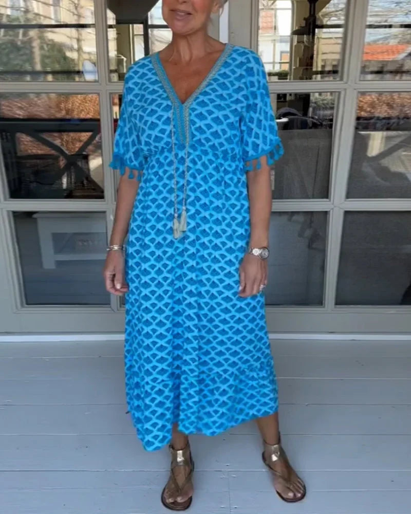 A woman walking near the coast in a flowing blue diamond-lattice print maxi dress with a tassel-trimmed neckline.
