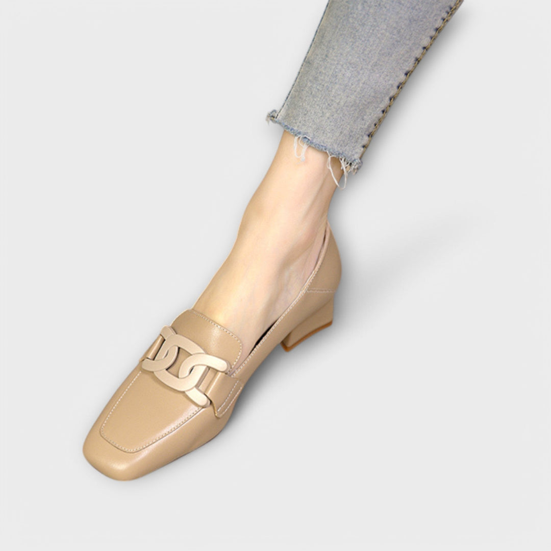 A pair of soft beige square-toe loafers with a chain-link ornament across the front.