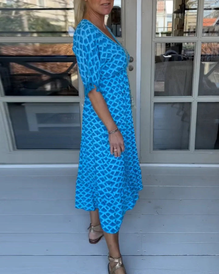 A woman walking near the coast in a flowing blue diamond-lattice print maxi dress with a tassel-trimmed neckline.