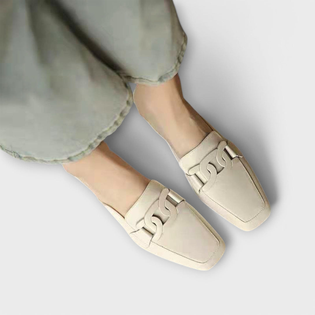 A pair of soft beige square-toe loafers with a chain-link ornament across the front.