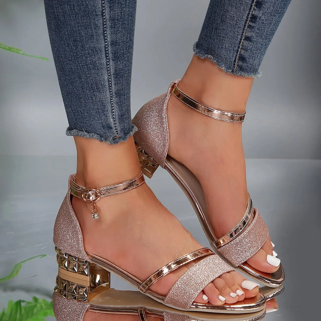 A woman wearing black sparkling block-heel sandals with a gold ankle strap and a crystal-set heel