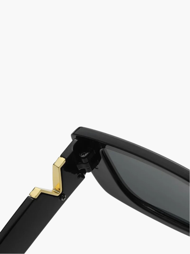 A woman at an outdoor event wearing chunky black rectangular retro sunglasses with a gold-tone accent.