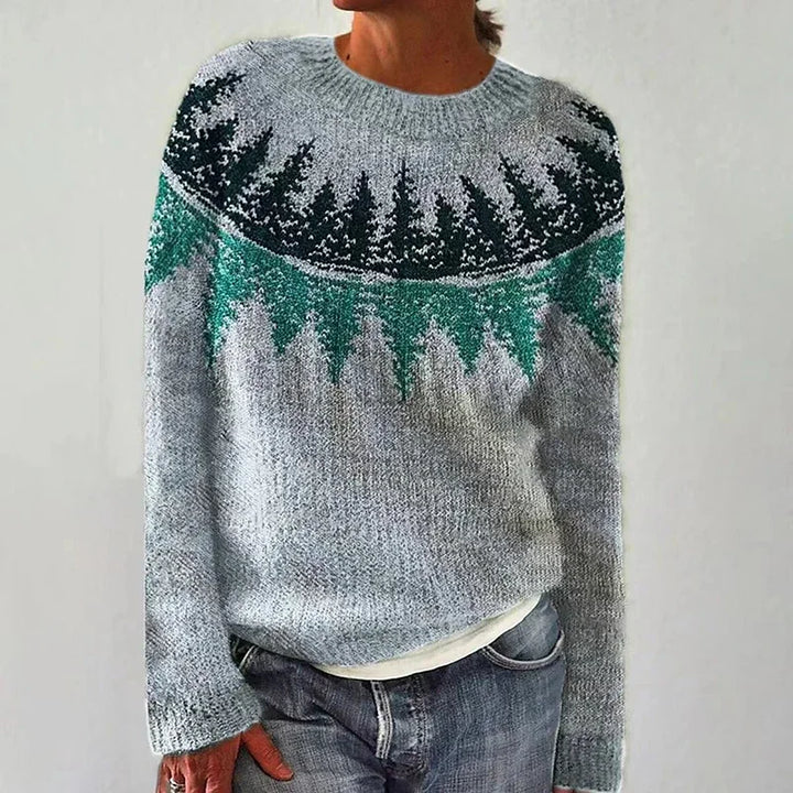 A woman wearing a sweater with a painterly pine forest print in pink, blue and grey, cosy and relaxed for fall.