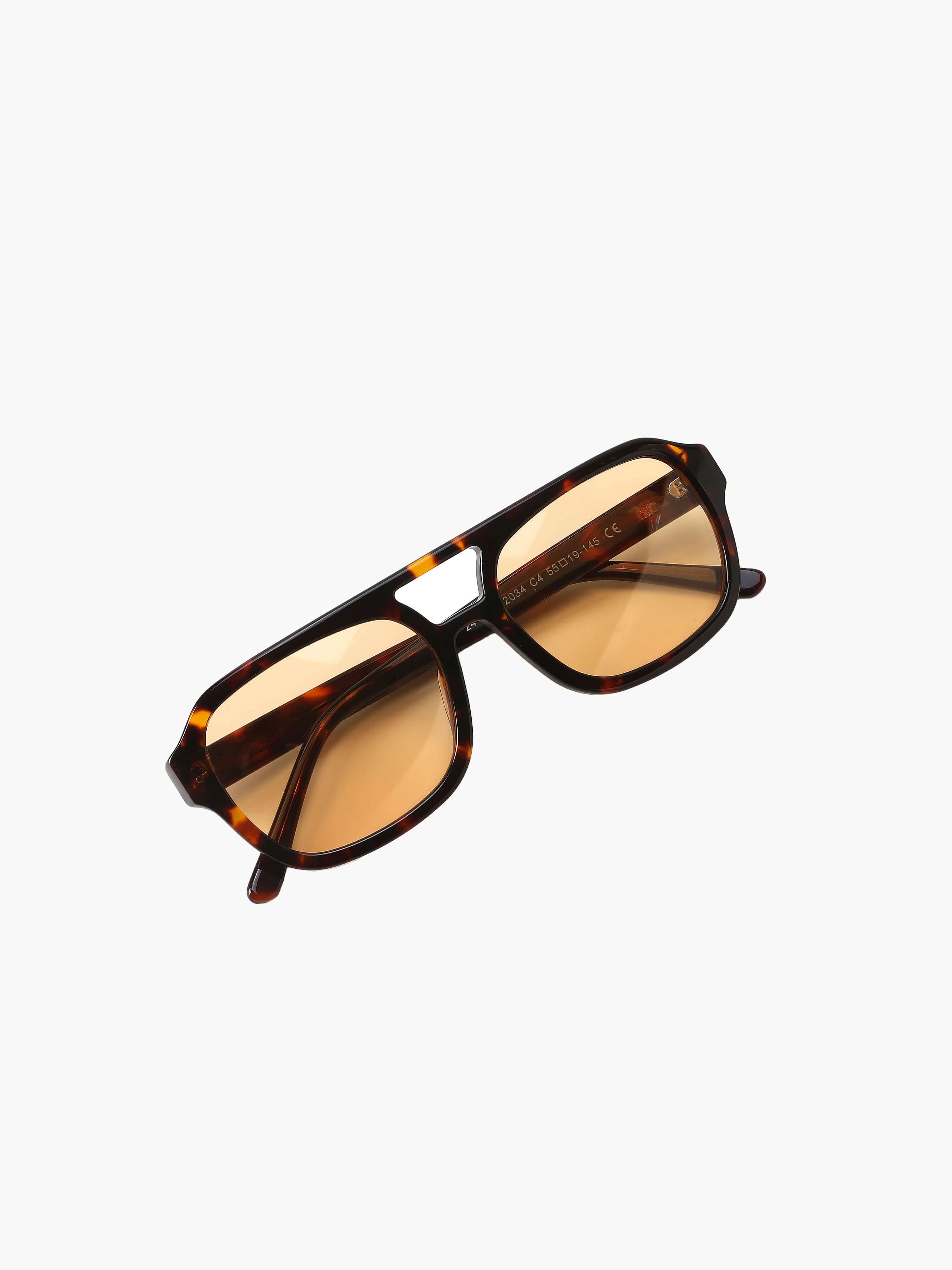 A woman wearing oversized tortoiseshell aviator sunglasses with green-tinted lenses on a sunny day outdoors.
