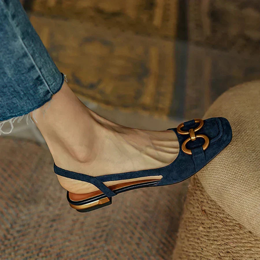 A woman wearing navy square-toe open-back flats with a small golden buckle detail.