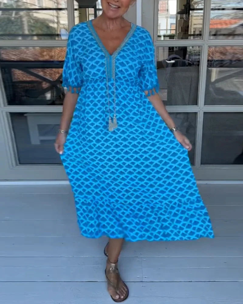 A woman walking near the coast in a flowing blue diamond-lattice print maxi dress with a tassel-trimmed neckline.