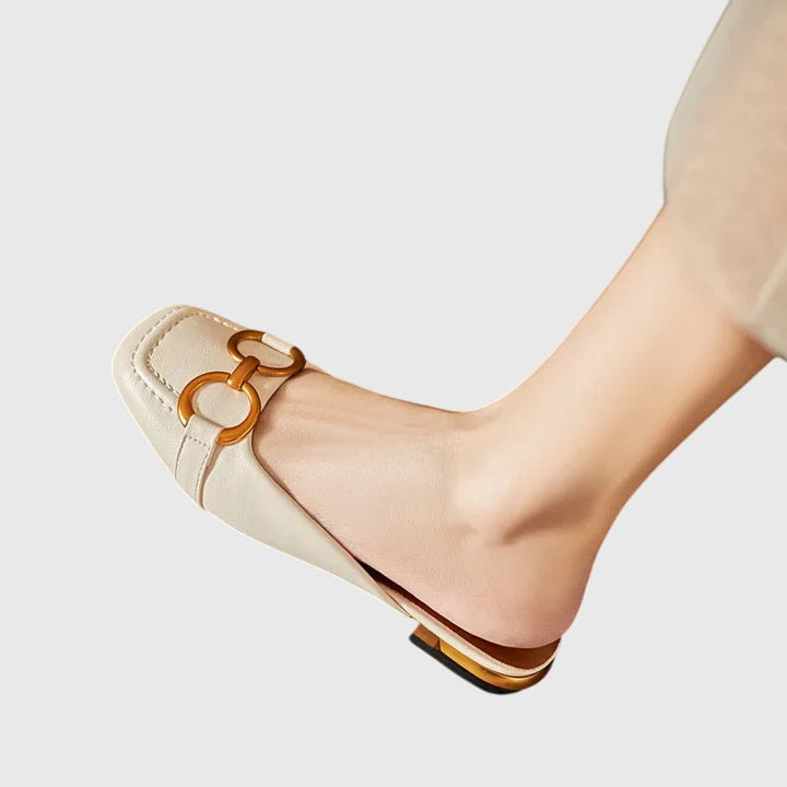 A cream closed-toe mule with a gold horsebit ring detail and a low heel.