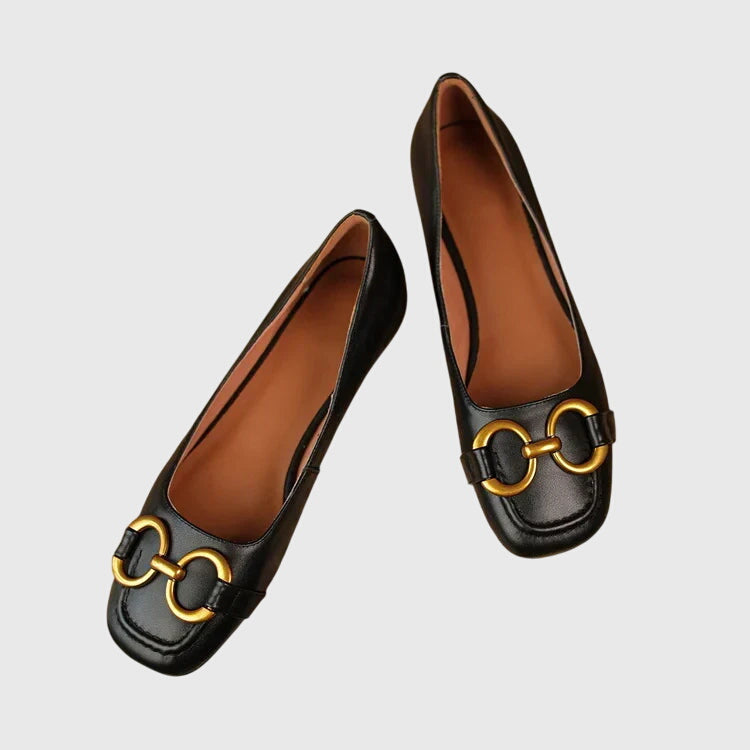A pair of black slip-on flats with a polished gold ring detail across the toe.