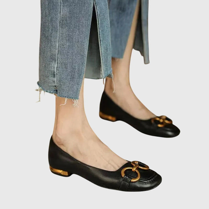 A pair of black slip-on flats with a polished gold ring detail across the toe.