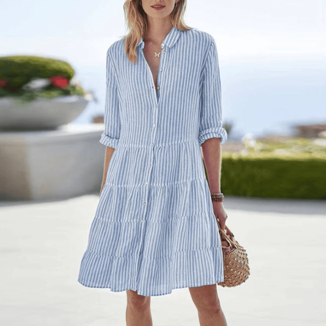 A woman wearing the Lori coastal stripe dress with a tiered skirt and button front, strolling by the sea.